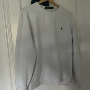 Vit sweatshirt från Polo Ralph Lauren - Vit sweatshirt från Polo Ralph Lauren med klassisk rund hals och broderad marinblå logga på bröstet. Tröjan är i bomull och har en relaxed fit med långa ärmar. Perfekt för en clean och stilren look.