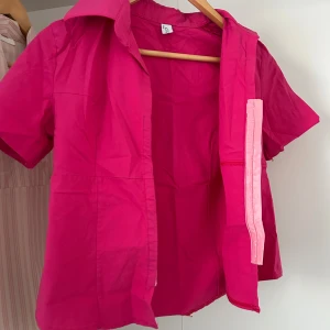 Chockrosa kortärmad skjorta - Chockrosa skjorta med korta ärmar och klassisk krage. Skjortan har en rak passform och stängs med hakar och är markerad med en ljusrosa kant. Perfekt för dig som vill sticka ut med färg. Materialet känns som bomull.