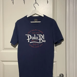 Marinblå t-shirt från Polo Ralph Lauren - Snygg marinblå t-shirt från Polo Ralph Lauren med tryck i vitt och rött framtill. Klassisk rund hals och korta ärmar. Tillverkad i mjuk bomull som känns skön mot huden. Perfekt för dig som gillar stilrena logotyper och ikoniska märken.