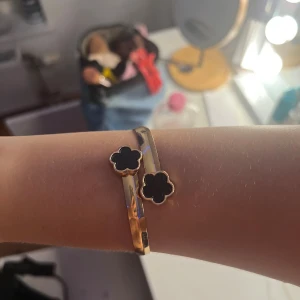 Guldigt armband Astrid & Agnes - Snyggt guldigt armband från Astrid & Agnes med två svarta blommor i klöverformad design. Armbandet har en stilren och modern look med blanka ytor och kontrasterande svarta detaljer. Perfekt för dig som gillar att sticka ut med accessoarer.