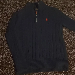 Mörkblå stickad tröja från Ralph Lauren - Mörkblå kabelstickad tröja från Ralph Lauren med halv dragkedja och röd broderad logga på bröstet. Tröjan har hög krage och långa ärmar, perfekt för kyliga dagar. Klassisk och stilren design som funkar till det mesta.