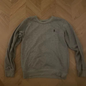 Grå sweatshirt från Ralph Lauren - Klassisk grå sweatshirt från Ralph Lauren med svart broderad logga på bröstet. Tröjan har rund halsringning, ribbade muddar och är tillverkad i mjuk bomullsmix. Den är sparsamt använd eftersom jag nu har storlek M.