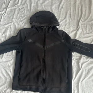 Svart hoodie från Nike med dragkedja - Svart hoodie från Nike i storlek S, med hel dragkedja och huva. Snygga svarta detaljer på bröstet och en ficka med dragkedja på ärmen. Tillverkad i mjukt bomullsmaterial, perfekt för chill eller träning. Passar dig som gillar sportig och enkel stil.