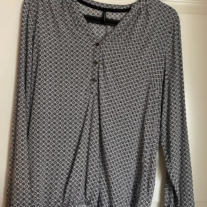 Svartvit mönstrad blus med knappar - Säljer en svartvit blus med mönster och långa ärmar. Blusen har v-ringning och dekorativa silver knappar framtill och vid armarna. Fin som vardagsblus