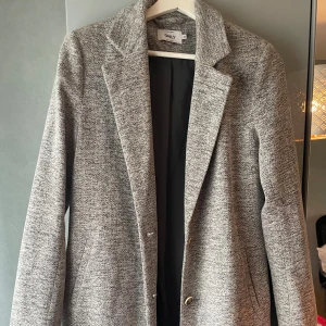 Grå kappa från ONLY  - Snygg gråmelerad kappa från ONLY med klassisk krage och två knappar framtill. Jackan har en rak passform.