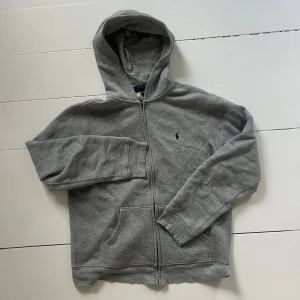 Grå fleece hoodie från Polo Ralph Lauren - Köpt på Kids Brand Store. Snygg grå hoodie från Polo Ralph Lauren i storlek XL. Tröjan har dragkedja, huva och två fickor framtill. Tillverkad i mjuk fleece och har det klassiska Polo-logot broderat på bröstet. Perfekt för chill dagar och enkel att matcha med jeans.