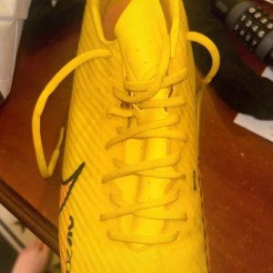 Nike Mercurial gula fotbollsskor - Säljer ett par Nike Mercurial fotbollsskor i en riktigt snygg gul färg med rosa och svarta detaljer. Skorna har snörning och är tillverkade i syntetmaterial för bästa grepp och känsla på planen. Perfekta för dig som vill sticka ut på fotbollsplanen.