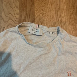 Ljusgrå t-shirt från Les Deux - Ljusgrå t-shirt från Les Deux med rund halsringning och diskret röd broderad II-logga på bröstet. Klassisk passform och tillverkad i mjuk bomull, perfekt för en clean och enkel stil.