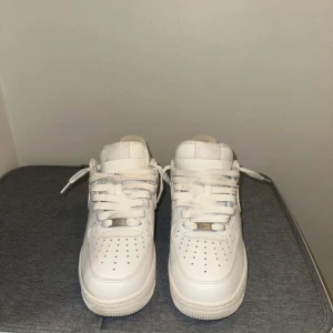 Nike Air Force 1 vita sneakers - Klassiska Nike Air Force 1 sneakers i helvitt skinn med rund tå och platt sula. Skorna har perforerade detaljer på tån, snörning och den ikoniska swoosh-loggan på sidorna. Perfekta för en clean och stilren look. 