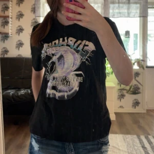 Svart t-shirt med draktryck - Cool svart t-shirt med stort tryck av en lila drake och text på framsidan. T-shirten har korta ärmar och en klassisk rund hals. Perfekt för dig som gillar edgy motiv och vill sticka ut lite extra.