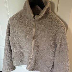 Beige teddyjacka från Lager157 - Bra skick! Lite defekt i kragen. Teddyjacka i beige från Lager157. Jackan har hög krage, dragkedja framtill och två stora fickor. Passformen är normal och materialet är fleece-liknande. 
