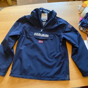 Blå vinterjacka från Napapijri - Snygg marinblå vinter/vind jacka från Napapijri med huva och halv dragkedja. Jackan har stor logga och norsk flagga framtill, samt fleecefoder på insidan för extra komfort. Praktisk sidodragkedja för enkel påtagning. Perfekt för blåsiga dagar. Jackan har tyvärr ett hål i armhålan, hålet går dock inte igenom fleece tyget. Därav påverkar det inte jackans värme. Jag har måtten på jackan ifall det behövs!!