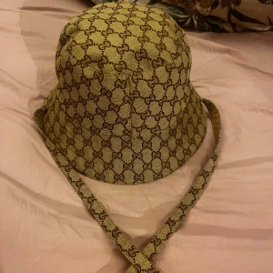 Beige bucket hat med snörning - Säljer en beige bucket hat med klassiskt GG-mönster i brunt. Hatten har bred brätte och långa snören med guldfärgade detaljer i ändarna. Materialet är kraftig bomull och insidan är fodrad för extra komfort. Perfekt för dig som vill sticka ut med lyxig streetstyle.