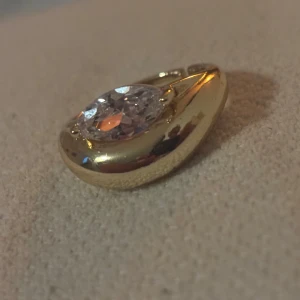 Guldfärgad ring med stor kristall - Snygg guldfärgad ring med en stor, ovalslipad klar kristall i mitten. Ringen har en chunky och modern design som verkligen drar blickarna till sig. Perfekt för dig som gillar statement-smycken och vill ha något som sticker ut. Tål vatten 