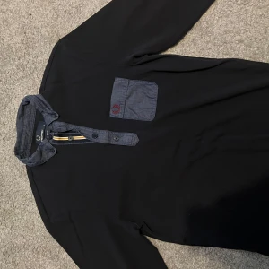 Svart henley från Fred Perry, slim fit - Svart henley-skjorta från Fred Perry i slim fit med kontrasterande blå krage och knappslå. Bröstficka i blått tyg med röd broderad logga. Snygga detaljer med gula och vita ränder vid knapparna. Perfekt för dig som gillar stilrena plagg.