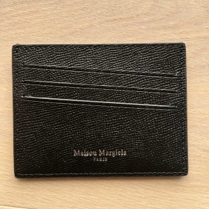 Maison Margiela svart korthållare i skinn - Minimalistisk svart korthållare från Maison Margiela i exklusivt präglat skinn. Har flera kortfack på båda sidor och de ikoniska vita sömmarna på baksidan. Kommer med original ask och dustbag. Perfekt för dig som vill ha stilren och lyxig accessoar.