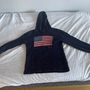 Mörkblå stickad hoodie från Ralph Lauren - Mörkblå stickad hoodie från Ralph Lauren med amerikansk flagga på bröstet och RL-broderi. Tröjan har huva, långa ärmar och bred ribbad mudd nertill. Perfekt för dig som vill ha en klassisk och snygg look med streetvibe.