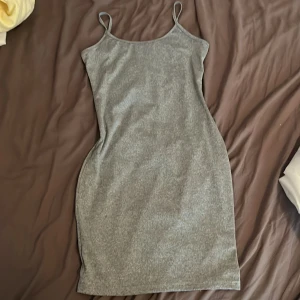 Grå  kortklänning - En enkel och stilren grå kortklänning med smala axelband. Klänningen har en tight passform och är ärmlös, perfekt för varma dagar eller att styla med en jacka. Materialet känns mjukt och bekvämt mot huden.
