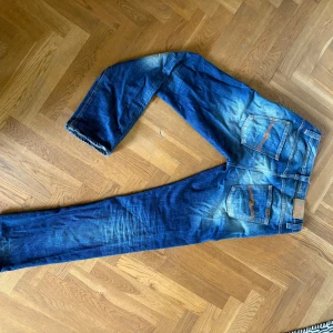 Nudie Jeans blå jeansbyxor - Säljer ett par klassiska blå jeans från Nudie Jeans med snyggt slitna detaljer och kontrastsömmar. Jeansen är tillverkade i 100% ekologisk bomull och har raka ben med normal passform. Bakfickorna har Nudie Jeans signaturbroderi.