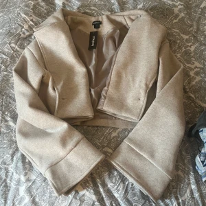 Beige croppad kappa från Boohoo - Säljer en beige croppad kappa från Boohoo med breda slag, lång ärm och en knapp framtill. Jackan har fickor och är fodrad för extra komfort. Perfekt för dig som vill ha en stilren och trendig look. Ny med prislapp. Storlek 38 men skulle säga att deras storlekar är alltid mindre.