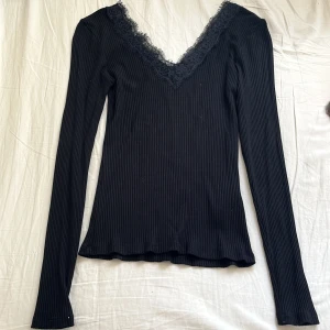 Svart ribbad topp med spets - Svart långärmad ribbad topp med djup v-ringning och snygg spetskant runt halsen. Toppen är figurnära och har en stilren look som passar perfekt till jeans eller kjol. Materialet är mjukt och stretchigt för en bekväm känsla 🖤