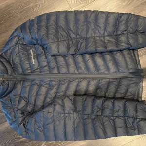 Peak Performance dunjacka  - Säljer en mörkblå dunjacka från Peak Performance med quiltad design och hel dragkedja framtill. Jackan är lätt och smidig, perfekt för kyliga dagar. Klassisk passform med lång ärm och logga på bröstet. 