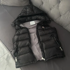 Svart dunväst från Moncler med huva - Snygg svart dunväst från Moncler med avtagbar huva och två dragkedjeförsedda fickor. Västen har quiltad design och klassisk Moncler-logga på bröstet. Perfekt för lager-på-lager och håller dig varm med stil.