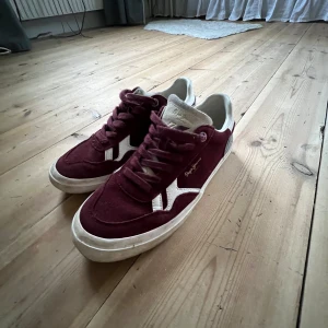 Vinröda sneakers från Pepe Jeans - Snygga vinröda sneakers från Pepe Jeans med vit sula och vita detaljer på sidan. Skorna har snörning och är tillverkade i mocka för en trendig look. Perfekta för dig som vill sticka ut med stilrena och bekväma skor.