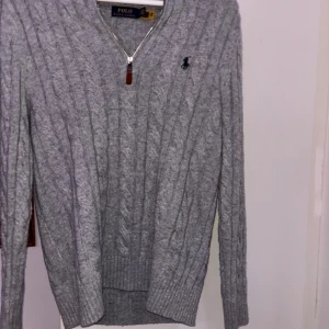 Grå kabelstickad tröja Polo Ralph Lauren - Kabelstickad grå tröja från Polo Ralph Lauren med half zip och klassisk broderad logga på bröstet. Långärmad modell i mjukt stickat material, perfekt för lager-på-lager. Snygg ribbad kant vid ärmslut och nederkant.