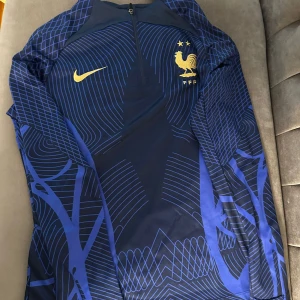 Frankrike Nike långärmad fotbollströja S - Exklusiv långärmad Frankrike-tröja från Nike i mörkblått med tunna blå linjer och guldiga detaljer. Broderad tupp och Nike-logga på bröstet. Materialet är lätt och andas, perfekt för träning eller match. Halv dragkedja framtill för extra stil.