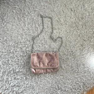 Rosa axelväska i sammet med kedja - Säljer en liten axelväska i ljusrosa sammet med snygg kedjerem och dekorativa kedjedetaljer runt kanterna. Väskan har ett mjukt, lyxigt tyg och stängs med lock. Perfekt storlek för det viktigaste och ger en cool touch till outfiten.