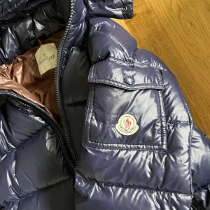 Mörkblå Moncler maya - Säljer en mörkblå pufferjacka från Moncler med glansig finish och huva. Jackan har dragkedja framtill, två sidofickor med dragkedja och en ficka med knapp på ärmen med Moncler-logga. Insidan är brun och jackan är fodrad för extra värme. Går att scanna