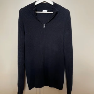 Mörkblå stickad half zip från John Henric - Mörkblå stickad tröja från John Henric med ribbad struktur och half zip-dragkedja vid halsen. Tröjan har lång ärm och hög krage, perfekt för kyligare dagar. Enkel och stilren design som funkar till många outfits.