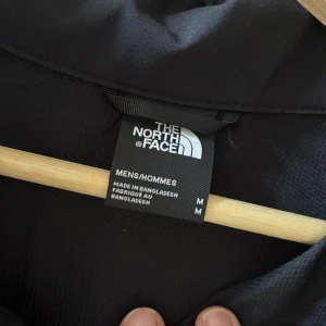 Svart vindjacka från The North Face M - Svart vindjacka från The North Face i storlek M. Jackan har dragkedja framtill, tre fickor med dragkedja och logga på bröstet och ryggen. Perfekt för blåsiga dagar och enkel att matcha med allt. Snygg och stilren design med hög krage. Frakt betalas av köparen