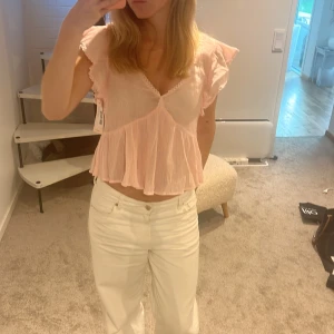 Ljusrosa volangblus med v-ringning - Supersöt ljusrosa blus med v-ringning och volangdetaljer. Blusen är croppad och har korta, vågiga ärmar samt spetskant vid halsen. Luftigt material som ger en romantisk vibe, perfekt för varma dagar. Helt oanvänd, lappar sitter kvar