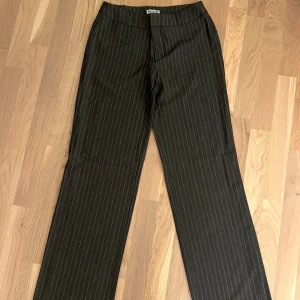 Svarta kostymbyxor med ränder från MadLady - Svarta kostymbyxor från MadLady med tunna vita ränder. Byxorna har liten bootcut och låg midja. Stretchigt och bekvämt material.