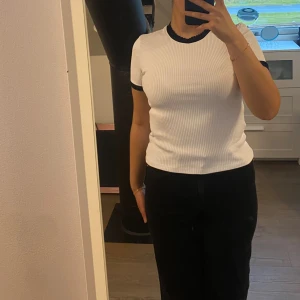 Vit ribbad t-shirt med svarta detaljer - Snygg vit ribbad t-shirt med svarta kanter vid hals och ärmslut. Modellen är kortärmad och har en tight passform som framhäver figuren. Perfekt för dig som gillar en enkel men stilren look.