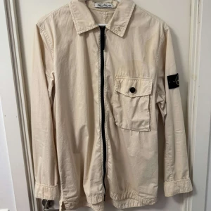 Beige overshirt från Stone Island - Snygg beige overshirt från Stone Island med klassisk krage, bröstficka med knapp och ikonisk patch på ärmen. Jackan har dragkedja framtill och är tillverkad i bomull, perfekt för lager-på-lager-stil. Passar dig som gillar clean och stilren look. L passar M