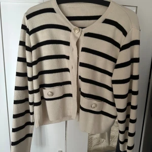 Randig beige kofta med guldknappar - Snygg beige kofta med svarta ränder och guldiga knappar framtill. Koftan har två små fickor med dekorativa knappar och är långärmad. Perfekt för dig som gillar en klassisk men trendig look. Materialet känns mjukt och stickat.