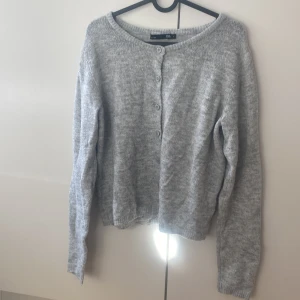 Grå stickad kofta från 157 - Mysig grå stickad kofta från 157 med rund hals och knappar framtill. Koftan har långärmade ärmar och ribbade muddar vid hals, ärmslut och nederkant. Perfekt för att hålla sig varm under kyliga dagar.