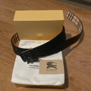 Burberry rutig skinnbälte med B-spänne - Säljer ett snyggt Burberry bälte i skinn med klassiskt rutigt mönster i beige, brun och svart. Bältet har ett stilrent metallspänne med Burberrys ikoniska B-logga. Perfekt accessoar för att lyfta din outfit med lyxig känsla och tidlös design.