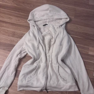 Vit hoodie med fuskpälsfoder FB Sister - Mysig vit hoodie från FB Sister med fluffigt fuskpälsfoder på insidan. Tröjan har dragkedja, två fickor fram och justerbar huva med snören. Perfekt för kyliga dagar när du vill vara både varm och snygg.