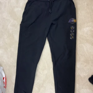 Lakers x Hugo Boss byxor - Svarta mjukisbyxor från Hugo Boss NBA-kollektion med Los Angeles Lakers-logga och BOSS-tryck i lila och gult på benet. Byxorna har snörning i midjan och är tillverkade i mjukt bomullsmaterial. Perfekta för chill eller sportiga dagar. Äkta!