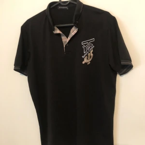 Svart Burberry t-shirt XL med tryck - Svart t-shirt från Burberry i storlek XL med klassiskt rutigt tyg vid kragen och snyggt Burberry London-tryck på bröstet. T-shirten har korta ärmar och en normal passform. Perfekt för dig som gillar stilrena designer med en touch av lyx. Xl i barnstorlek och m i vuxenstorlek ver ej om den är äkta