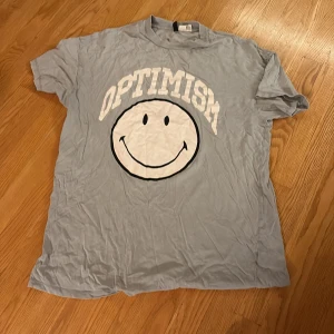 Grå t-shirt Smiley x Divided S - Oversized ljusgrå t-shirt från Smiley x Divided i storlek S. Stor vit smiley och texten 'OPTIMISM' tryckt på bröstet. Klassisk rund hals och korta ärmar. Skön och avslappnad passform, perfekt för en chill look.