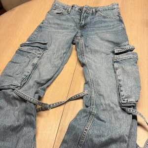 Cargo Jeans stora fickor från Zara, strl 34 - Säljer ett par Jeans cargo från Zara i storlek 34. Byxorna har flera stora fickor på benen, råa och slitna benslut samt banddetaljer. Tillverkade i klassiskt jeansmaterial med loose passform och cool streetstil.