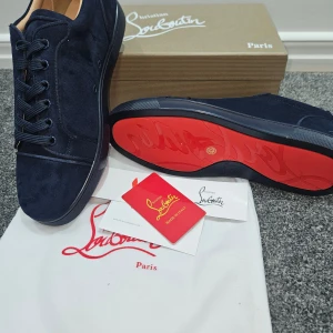 Mörkblå sneakers från Christian Louboutin - Snygga mörkblå sneakers från Christian Louboutin i mjuk mocka med klassisk röd sula. Skorna har rund tå, snörning och diskreta sömdetaljer. Insidan är beige med guldfärgad logga. Perfekt för dig som gillar exklusiv stil och vill sticka ut med ikoniska detaljer.