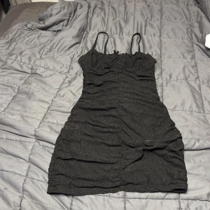 Svart kort klänning från H&M  - Svart kort klänning från H&M Divided med smala axelband och lätt rynkad struktur. Klänningen är figurnära och har en enkel, stilren design som passar perfekt för en trendig look. Materialet är mjukt och stretchigt för en bekväm passform.