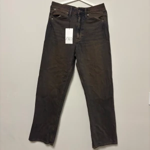 Zara - Klassiska slim fit jeans i mörkbrun ton med vintage-look. Rak passform med lätt stretch för komfort hela dagen. Perfekt för vardagsoutfits.  Nyckelfunktioner: •  Mid rise midja för bekväm sittning. •  Slim genom benet, avslutas rak vid foten. •  Mjukt denim med elastan för rörelsefrihet. •  Fickor framtill och bak, dragkedja och knapp. •  Lätt wash-effekt för sliten stil. 
