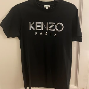 Svart Kenzo Paris t-shirt XS - Svart t-shirt från Kenzo Paris i storlek XS. Klassisk modell med rund hals och korta ärmar. Framsidan har ett stort, grafiskt Kenzo Paris-tryck i vitt. Materialet är mjuk bomull.